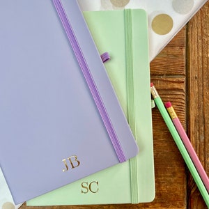May include: Two notebooks with elastic closures, one lavender and one light green, with gold lettering. The lavender notebook has the initials "JB" and the light green notebook has the initials "SC".