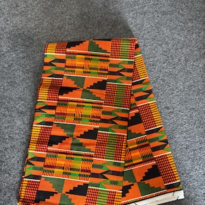 May include: Folded orange, green, and black patterned fabric with a white border. The fabric has a geometric design with the text "100% COTTON" printed on the border. The fabric is folded over on a gray surface.