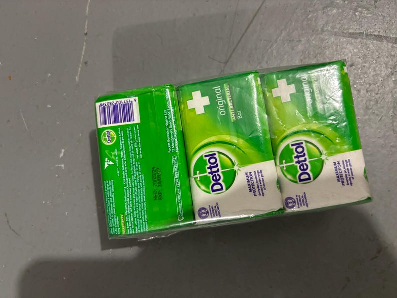 May include: A three-pack of Dettol Original Anti-Bacterial soap bars wrapped in clear plastic. Each green and white bar features the Dettol logo, a white cross, and the text Maximum Protection. The packaging includes a barcode and ingredient list.