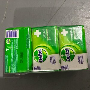 May include: A three-pack of Dettol Original anti-bacterial soap bars wrapped in clear plastic. The packaging is bright green and white, featuring the Dettol logo, a white cross symbol, and text detailing product information and ingredients.