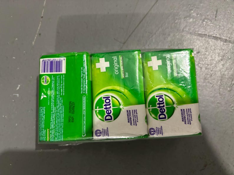 May include: A three-pack of Dettol Original anti-bacterial soap bars wrapped in clear plastic. The packaging is bright green and white, featuring the Dettol logo, a white cross symbol, and text detailing product information and ingredients.