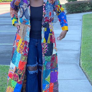 Puede incluir: Un abrigo largo de patchwork en una variedad de colores, incluyendo rojo, azul, amarillo y morado. El abrigo está hecho de diferentes patrones y texturas de tela. La persona lleva un top negro y vaqueros azules.