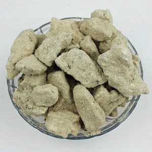 May include: A clear glass bowl filled with light beige, irregularly shaped, rock-like objects. The objects have a rough, textured surface and are piled together. The bowl sits on a white surface.