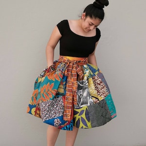 May include: A woman wearing a black top and a colourful patchwork skirt with a yellow belt. The skirt is made of different fabrics with various patterns and colours.
