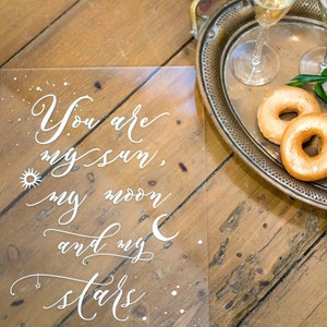 May include: A clear acrylic sign with the text "You are my sun, my moon and my stars" written in white calligraphy. The sign is on a wooden surface with a silver tray and donuts in the background.