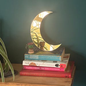 Peut inclure: Un miroir en forme de croissant de lune doré est posé sur une pile de livres sur une étagère en bois. Les livres ont des couvertures colorées et des titres tels que "ZEN", "notes from a small island" et "THE THIRD POLICEMAN". Un petit objet décoratif est posé sur les livres.