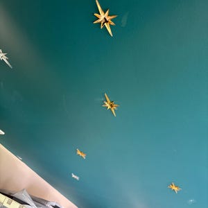 Könnte beinhalten: Eine blaugrüne Decke, die mit mehreren goldfarbenen, sternförmigen Wandaufklebern verziert ist. Die Sterne haben ein vielzackiges Design mit scharfen, länglichen Strahlen. Die Aufkleber sind in einem verstreuten Muster auf der Fläche angeordnet.