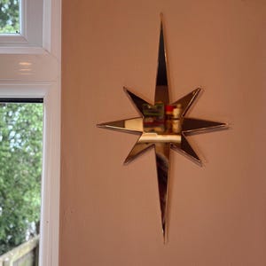 Op de afbeelding: Een gouden sterrenspiegel is bevestigd aan een perzikmuur. De spiegel heeft een centraal reflecterend oppervlak en acht puntige stralen. Deze decoratieve wanddecoratie is een stijlvolle toevoeging.