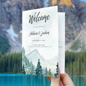 May include: A wedding program with a watercolor illustration of a mountain range with trees. The text reads "Welcome to the wedding of Addison & Jackson, September 11, 2021, Laurel Ridge Lodge, 1234 Laurel Ridge Road, Greensburg, CO."