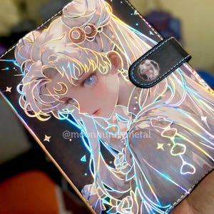 May include: A black and iridescent planner cover featuring a detailed anime-style illustration of a woman with long white hair and blue eyes. The cover has a black strap with a decorative emblem and the text "@moonbunny metal".
