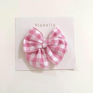 Puede incluir: Un lazo para el pelo de cuadros rosas y blancos sobre una tarjeta blanca. El lazo es de tela y tiene forma de lazo clásico. La tarjeta tiene el nombre "Piabella" impreso.
