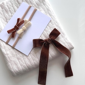May include: A set of three velvet hair bows in brown and cream colours. The bows are tied with a simple knot and are attached to elastic headbands. The headbands are shown on a white knitted blanket.