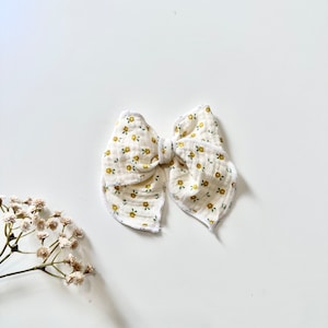 Könnte beinhalten: Eine weiße Schleife aus Stoff mit gelbem Blumenmuster. Die Schleife hat einen gewellten Rand und ist mit einem einfachen Knoten gebunden.