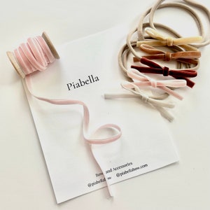 Puede incluir: Un carrete de cinta de terciopelo rosa claro y varios lazos pequeños de terciopelo en varios colores, incluyendo rosa, marrón y beige, se muestran sobre una superficie blanca. Los lazos están sujetos a diademas beige. El texto "Piabella" está impreso en una tarjeta blanca.