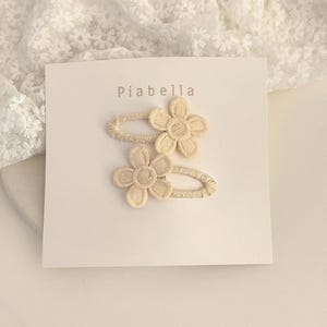 Puede incluir: Dos pinzas para el pelo con forma de flor color crema sobre una tarjeta blanca con el nombre "Piabella" impreso en la parte superior. Las pinzas están hechas de un material tejido y tienen un diseño floral. El fondo es una tela de encaje blanco.