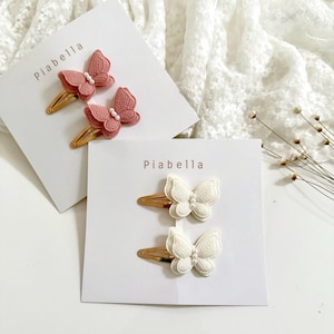 May include: Two sets of butterfly hair clips. One set features two pink butterfly clips with gold-colored metal clasps and small white beads. The other set has two white butterfly clips with gold-colored metal clasps. The brand name "Piabella" is printed on the cards.