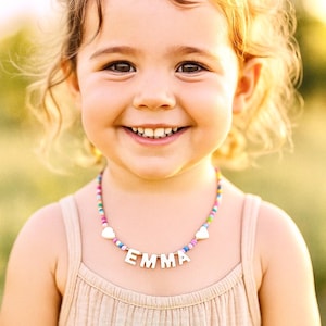 Puede incluir: Una niña sonriente lleva un collar de cuentas de colores con el nombre "EMMA" escrito con letras de madera. El collar tiene detalles en forma de corazón. La niña lleva una camiseta sin mangas beige.