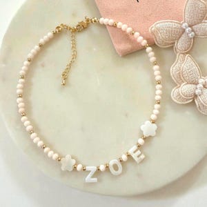 Puede incluir: Un delicado collar con el nombre "ZOE" escrito en letras blancas. El collar está hecho de pequeñas cuentas de color rosa claro y detalles dorados. También incluye dos dijes blancos con forma de flor.