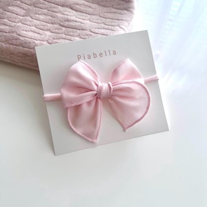 May include: A pink satin bow headband with the text "Piabella" on a white card.