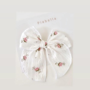 Puede incluir: Un lazo para el pelo de encaje blanco con detalles florales rosas. El lazo presenta un diseño calado y está adornado con pequeñas rosas rosas bordadas. El nombre de la marca "Piabella" es visible.