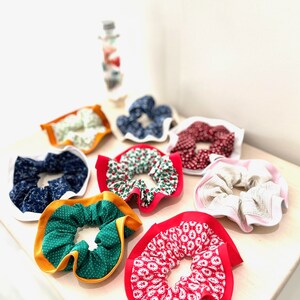 May include: A collection of handmade scrunchies in various colors and patterns. The scrunchies feature fabrics with polka dots, floral prints, and solid colors, with contrasting trim. The scrunchies are arranged on a light-colored surface.