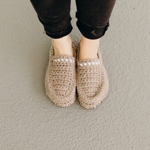 Puede incluir: Un par de zapatillas de crochet beige con ribete blanco. Las zapatillas están siendo usadas por una persona de pie sobre una superficie de concreto gris.