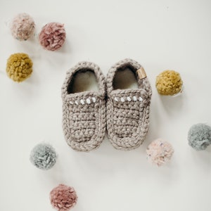 May include: A pair of handmade, gray crochet baby slippers with white stitching detail. The slippers are surrounded by colorful, fluffy pom-poms in shades of pink, yellow, and gray, creating a cozy and playful aesthetic.