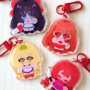 May include: Four acrylic keychains featuring stylised cartoon characters with red lobster-claw clasps. One character has dark hair and a chainsaw, one has orange hair, one has blonde hair with a frog, and one has red hair holding a white knife.