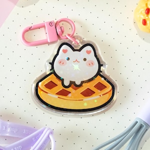 May include: A clear acrylic keychain with a cartoon cat on a waffle. The cat is white with pink accents, and the waffle is yellow and brown. The keychain has a pink clasp. The overall design is cute and whimsical.