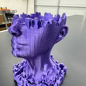 May include: A purple 3D printed sculpture of a human head with a cityscape design. The head is tilted to the side and the cityscape is visible on the top and sides of the head.