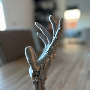 Könnte beinhalten: Eine silberne, 3D-gedruckte Roboterhand-Skulptur mit detailliertem Design. Die Hand ist in einer Geste des Ausstreckens posiert, mit ausgestreckten und leicht gekrümmten Fingern. Die Skulptur ist auf einem sechseckigen Sockel montiert.