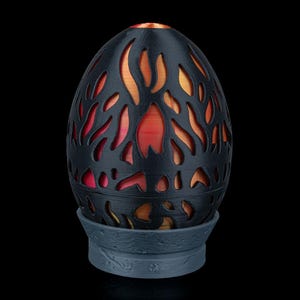 May include: A black, egg-shaped decorative lamp with flame-like cutouts. The lamp sits on a grey base and emits a warm orange and red glow. The lamp's design creates a unique ambiance.
