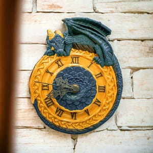 May include: A decorative wall clock featuring a blue and gold dragon perched atop. The clock face has Roman numerals and a floral design in the center. The clock is mounted on a white brick wall.