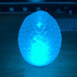 Peut inclure: Une lampe blanche en forme d'œuf avec une surface texturée ressemblant à des écailles de dragon est éclairée par une lumière bleue.