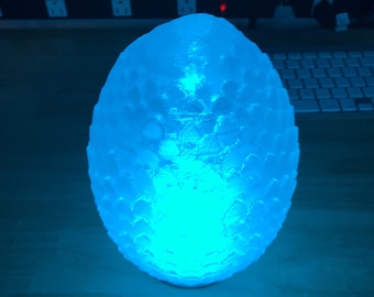 May include: A blue light illuminates a white, egg-shaped lamp with a textured surface resembling dragon scales.