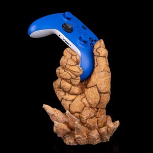 May include: A blue and white video game controller is held by a brown, textured hand-shaped stand. The stand has a rocky appearance, and the controller is a vibrant blue color.