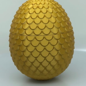Könnte beinhalten: Eine goldene Dracheneier-Schmuckschatulle mit einer strukturierten, schuppigen Oberfläche. Die Schachtel ist geöffnet und zeigt das Innere.