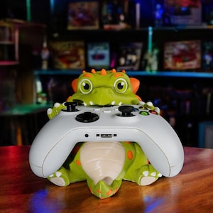 Op de afbeelding: Een groen en oranje dinosaurusvormige controllerhouder. De houder is ontworpen om een witte gamecontroller te ondersteunen. De dinosaurus heeft grote ogen en oranje stekels. De controller heeft zwarte knoppen en joysticks.