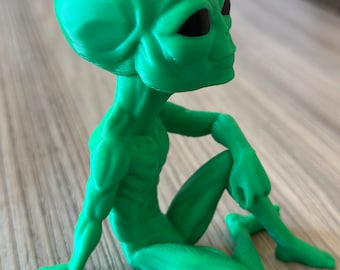 May include: A green alien figurine sitting with its legs crossed and one arm resting on its knee. The alien has a large head with black eyes and a slight smile.