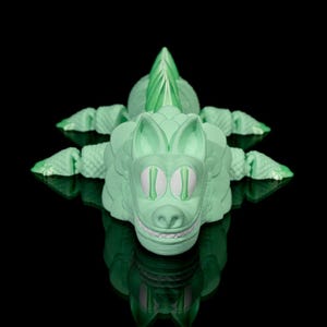 May include: A light green alligator figurine with a cartoonish design. The alligator has a smiling mouth, white teeth, and white eyes. The figurine is on a black surface, and its reflection is visible.