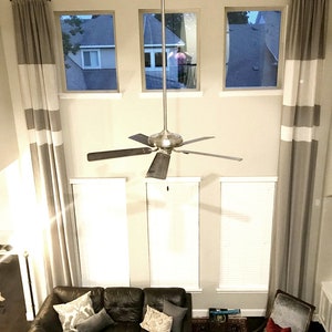 May include: Interior shot of a living room with tall windows and gray and white striped curtains. A dark brown leather sofa is in the foreground, with decorative pillows. A ceiling fan hangs from the ceiling.