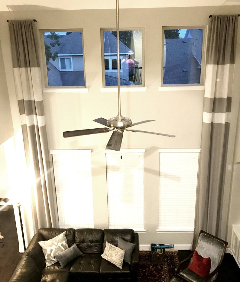 May include: Interior shot of a living room with tall windows and gray and white striped curtains. A dark brown leather sofa is in the foreground, with decorative pillows. A ceiling fan hangs from the ceiling.