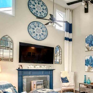 May include: A living room with a blue and white color scheme. The room features a fireplace, a large TV, and decorative wall art. Blue floral paintings and round wall hangings add to the decor. White curtains with blue stripes and a ceiling fan are also visible.