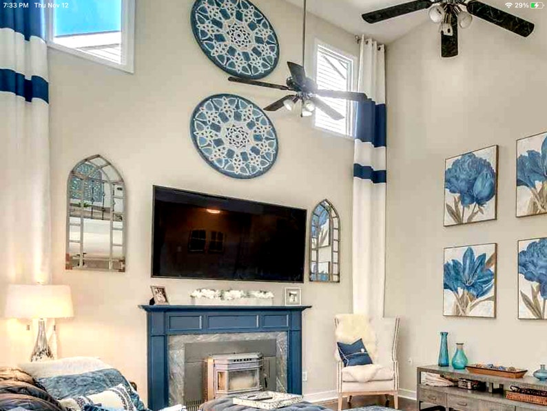 May include: A living room with a blue and white color scheme. The room features a fireplace, a large TV, and decorative wall art. Blue floral paintings and round wall hangings add to the decor. White curtains with blue stripes and a ceiling fan are also visible.