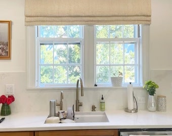 May include: A kitchen scene featuring a window with a beige patterned Roman shade. The window overlooks a lush green outdoor view. A white countertop with a sink, faucet, and various kitchen items is visible. Wooden cabinets are below the countertop.