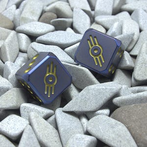May include: Two blue dice with gold markings, each featuring a stylized design, rest on a bed of gray stones. The dice have rounded edges and gold-colored dots indicating the numbers. The dice are a deep blue color.