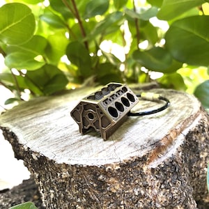 May include: A bronze-colored miniature engine block pendant with a black cord. The detailed model sits on a weathered tree stump, surrounded by green foliage. The engine block features multiple cylinders and intricate design.