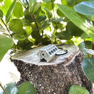 May include: A silver-colored miniature engine block pendant with a black cord. The pendant is resting on a tree stump, surrounded by green leaves. The engine block has multiple holes and a detailed design.