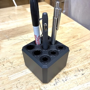 May include: A dark gray pen holder with six circular openings, holding a black Sharpie marker, a small gray pen, and a silver pen. The pen holder is a cube shape and sits on a wooden surface.