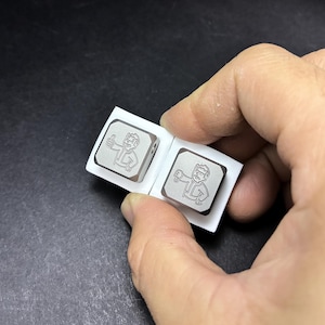 May include: Two square, silver-colored metal buttons with engraved cartoon figures. One figure gives a thumbs-up, the other a thumbs-down. The buttons are attached to a white paper backing, held by a hand.